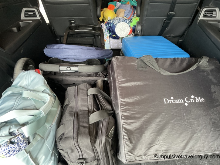 Rear cargo area of an SUV packed for a family road trip, with a teal duffel bag, black luggage, a folded navy stroller, a blue hard-shell suitcase, and a Dream On Me portable travel crib in a black carrying case, surrounded by children's toys and stuffed animals