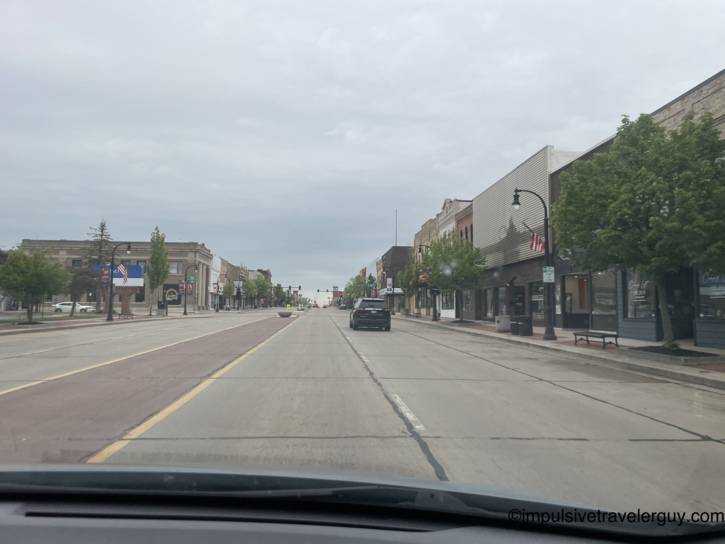 View through a car windshield of a quiet small-town main street lined with brick storefronts, American flags, street lamps, and a black SUV ahead under an overcast sky
