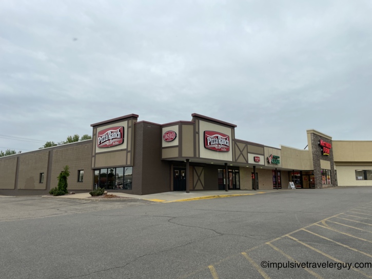 Exterior of a Pizza Ranch Buffet restaurant in a strip mall with a brown and tan facade, alongside Mattress Firm and Check Into Cash, with an empty parking lot in front under a cloudy sky