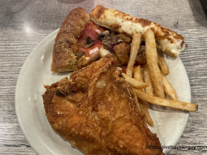 White plate of buffet food with a slice of supreme pizza, a cheese breadstick, fried chicken, french fries, and assorted toppings on a wood-grain table