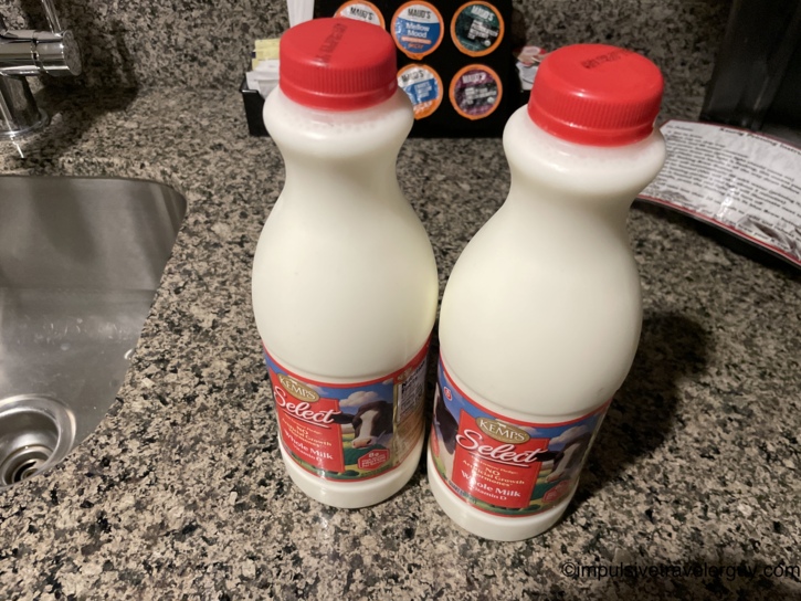 Two quart bottles of Kemps Select whole milk with red caps on a granite kitchen counter beside a sink, with Maud's coffee pods in the background