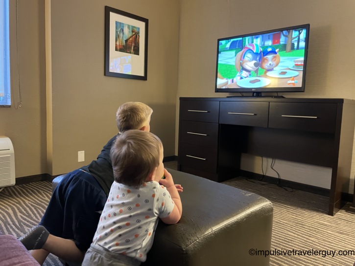 Two young boys sitting on an ottoman in a hotel room watching a PAW Patrol cartoon on a flat-screen television set atop a dark wood dresser