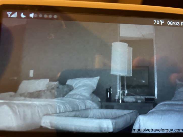 Blurry baby monitor screen showing a dark hotel room with two beds, a lamp, and a nightstand in night-vision mode, displaying 70 degrees Fahrenheit and 8:03 PM
