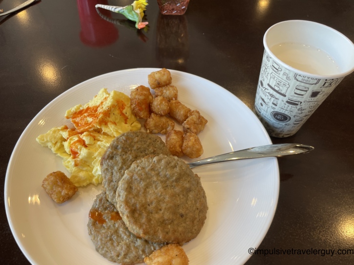 Breakfast plate with scrambled eggs topped with hot sauce, two sausage patties, and tater tots, next to a paper coffee cup on a dark table