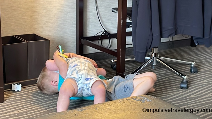 Two young children lying on patterned hotel carpet beneath a desk playing with toys, with an office chair and hanging clothes nearby