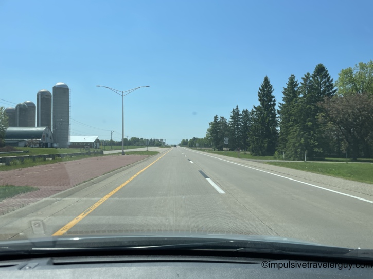 View through a car windshield of US-10 West, a divided rural highway under a clear blue sky, with farm silos and a barn on the left and evergreen trees on the right