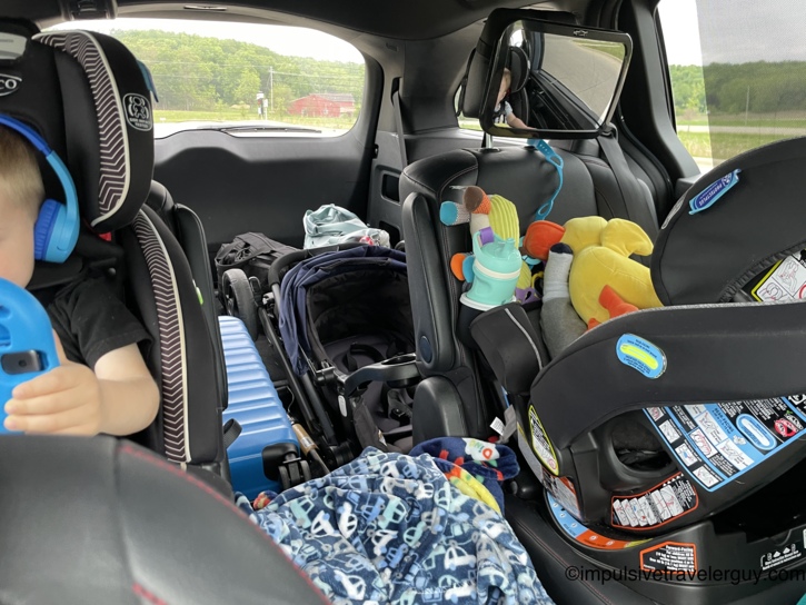 View from the back of a minivan showing a young boy in a car seat wearing blue headphones, a rear-facing infant car seat with attached toys and a teal sippy cup, and luggage packed in the middle including a blue suitcase and navy stroller, with a baby reflected in the overhead mirror