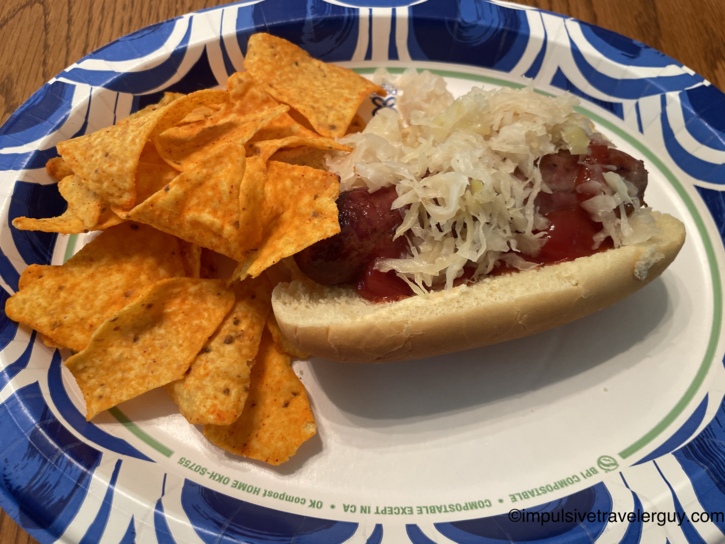 Paper plate with a grilled bratwurst in a bun topped with sauerkraut and ketchup, served with a side of Doritos on a blue and white patterned plate