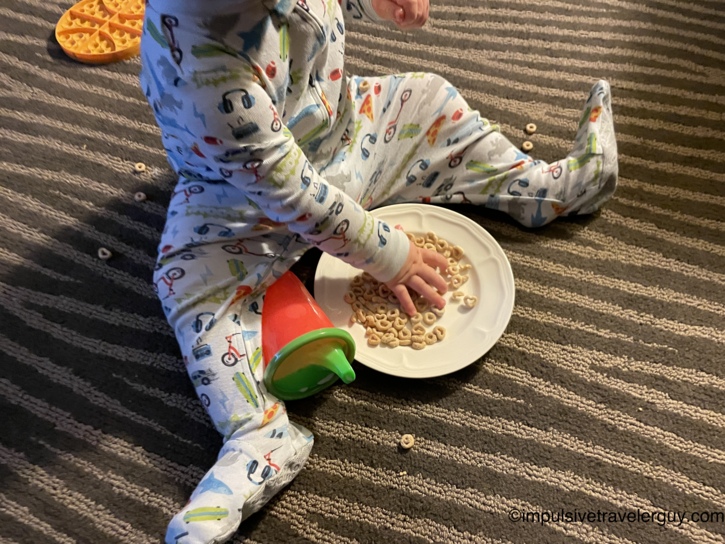 Baby in airplane-print footed pajamas sitting on patterned carpet eating Cheerios off a white plate, with an orange waffle teether and a green and red sippy cup nearby and stray cereal scattered around