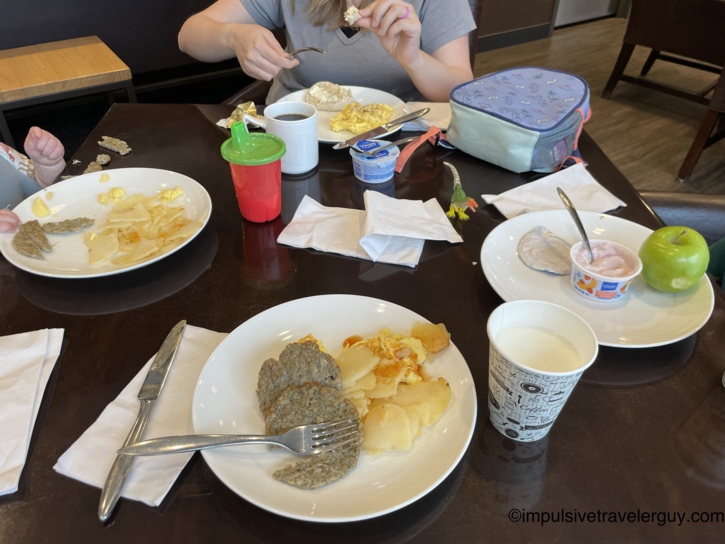 Hotel breakfast table with several white plates of scrambled eggs, sausage patties, and sliced potatoes, alongside yogurt cups, a green apple, coffee, a child's red sippy cup, and a lunch bag, with a woman eating in the background