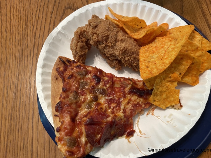 Paper plate with a slice of sausage and pepperoni pizza, fried chicken strips, and Doritos on a wood table