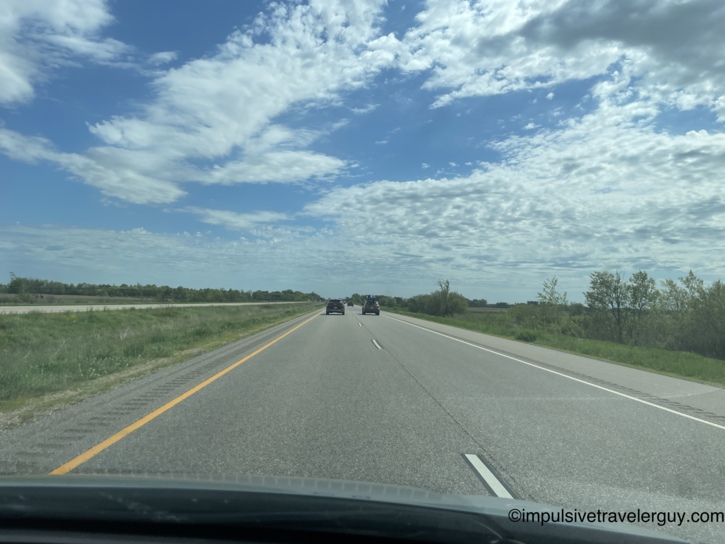 View through a car windshield of a divided highway under a blue sky with scattered clouds, vehicles ahead and green fields and trees lining both sides