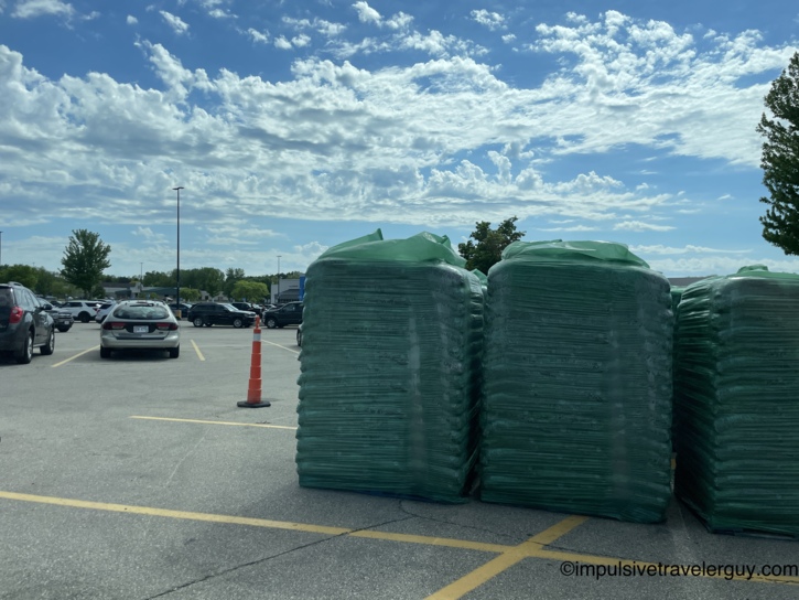 Large shrink-wrapped pallets of green-bagged mulch or soil stacked in a store parking lot, with parked cars, a traffic cone, and a partly cloudy sky