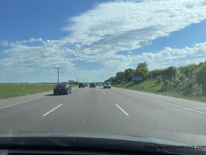 View through a car windshield of Interstate 39/90/94 South-East with traffic ahead, a blue interstate route sign on the right, and green embankments under a blue sky with clouds