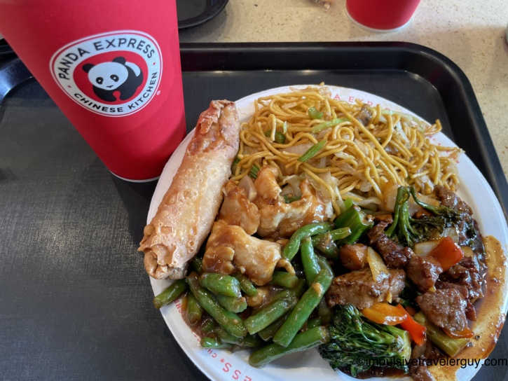 Panda Express meal on a black tray with chow mein, an egg roll, orange chicken with string beans, and Beijing beef with broccoli and peppers, beside a red Panda Express Chinese Kitchen drink cup