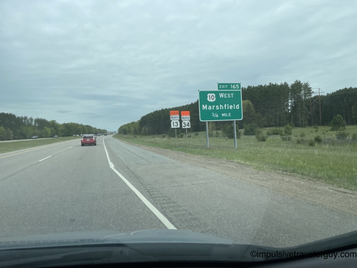View through a car windshield of a highway with a green Exit 165 sign for US-10 West toward Marshfield three-quarters of a mile ahead, alongside orange detour signs for North 13 and 34, under an overcast sky