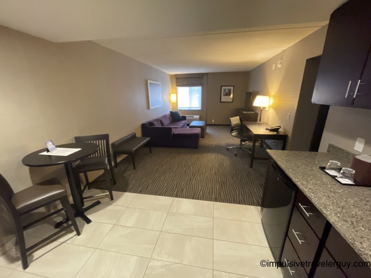 Hotel suite living area with a purple sofa and ottoman, a work desk with office chair, a high-top dining table with stools, a granite kitchenette counter with mini-fridge, and zebra-patterned carpet