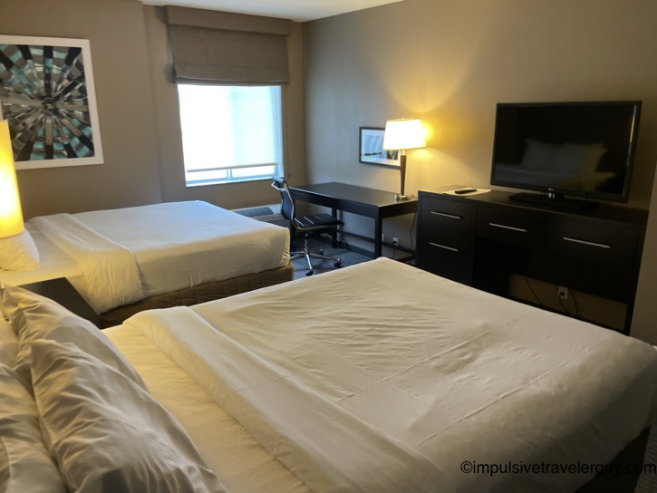 Hotel room with two queen beds in white linens, a dark wood desk and dresser, a flat-screen television, a curtained window, and abstract artwork on the wall
