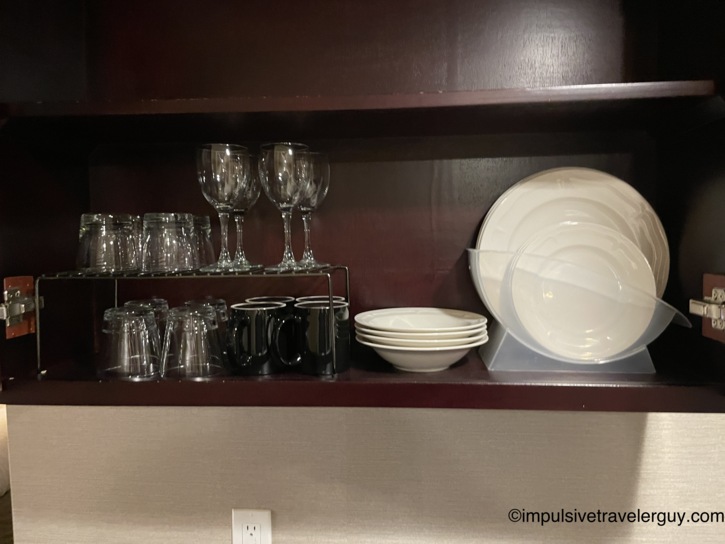 Open kitchenette cabinet stocked with wine glasses, drinking glasses, black coffee mugs, and white plates and bowls arranged on a wire rack and plastic stand against dark wood shelving