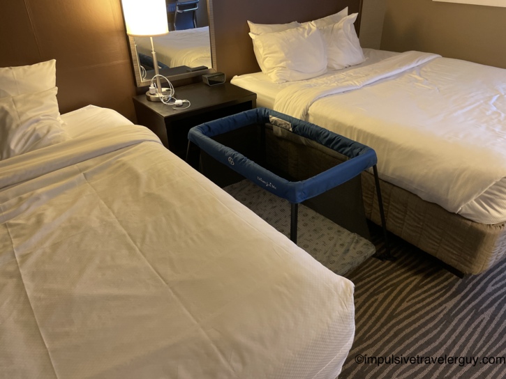 Hotel room with two beds and a blue Dream On Me portable travel crib set up on the floor between them, with a nightstand, lamp, mirror, and charging cables nearby