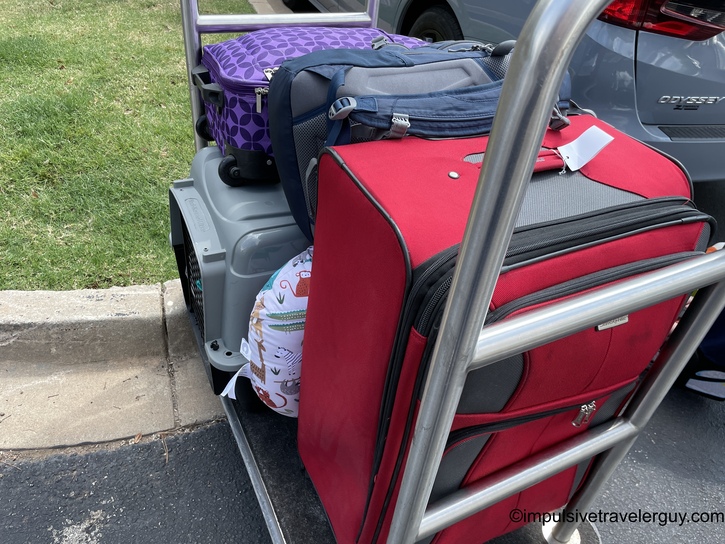 Hotel luggage cart loaded with red large suitcase, purple patterned bag, gray travel bag, and white diaper bag positioned at vehicle door