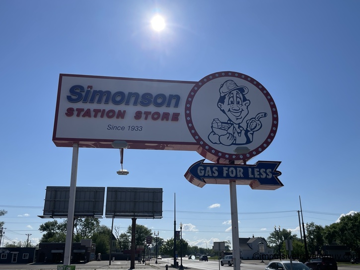 Vintage tall roadside sign reading "Simonson Station Store Since 1933" with retro cartoon attendant character logo and "Gas For Less" banner