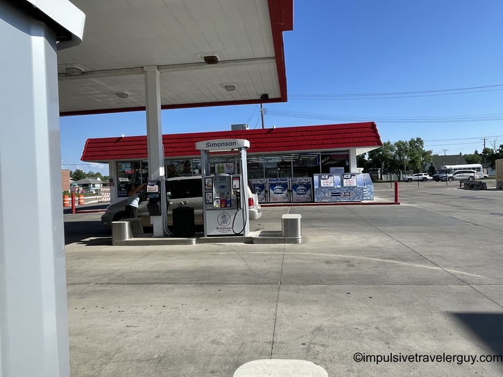 Gas station pump island with white overhead canopy and red-striped convenience store building displaying Simonson branding