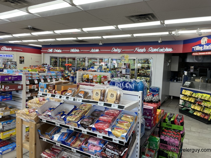 Convenience store interior with center aisle product displays, wall of refrigerated beverage cases, and red and blue "Filling Station" branding