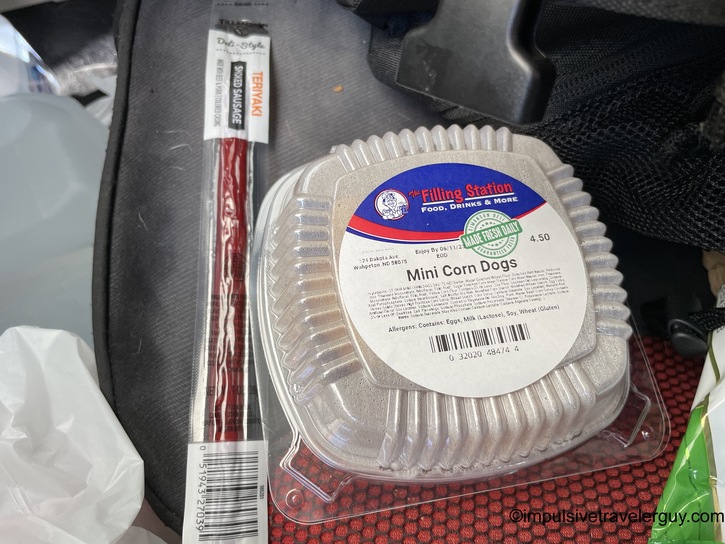 Clear plastic container of mini corn dogs and wrapped beef stick snack positioned on gray textured car seat