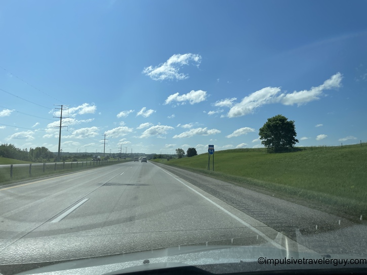 Rural divided highway from driver's perspective with single vehicle ahead under bright blue sky with scattered white clouds and power lines