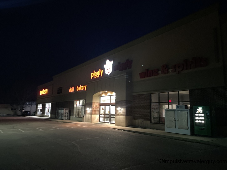 Night view of a strip mall storefront featuring a Piggly Wiggly grocery store with illuminated signage, deli and bakery sections visible, along with adjacent businesses in a parking lot