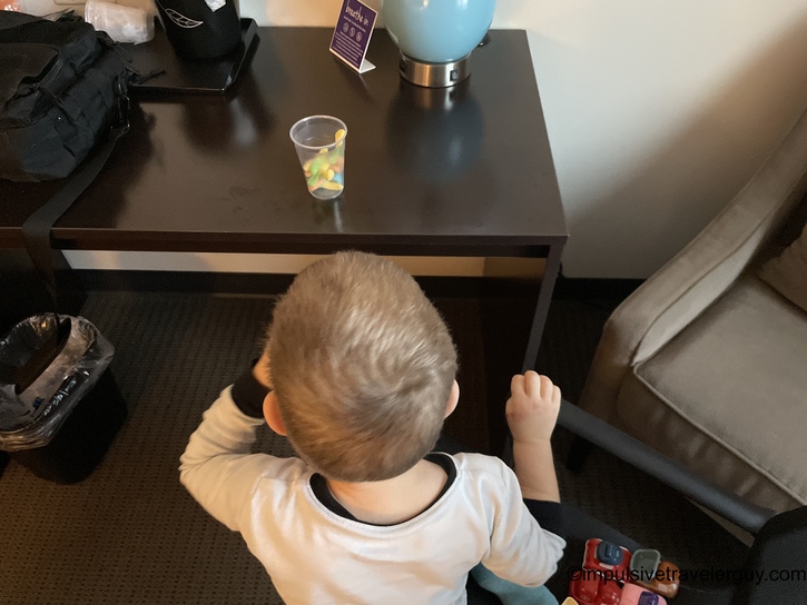 Young child in striped shirt sitting at hotel desk with cup of gummy snacks and toy vehicles visible