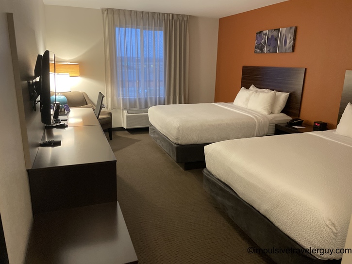 Modern hotel room interior with two queen beds, white linens, dark wood headboards, orange accent wall, desk area with TV and office chair, window with curtains