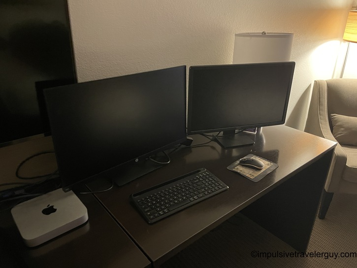 Hotel desk workspace setup with Mac Mini computer, dual monitors on stands, wireless keyboard and mouse on dark surface with lamp and chair