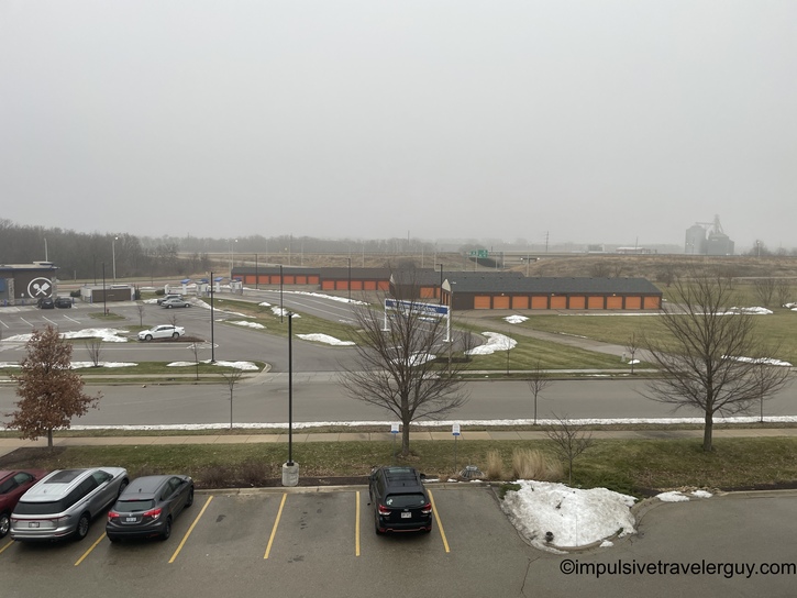 Overcast daytime view from hotel window showing Culver's restaurant, self-storage facility with orange doors, parking lot, bare trees, and grain elevator in foggy distance