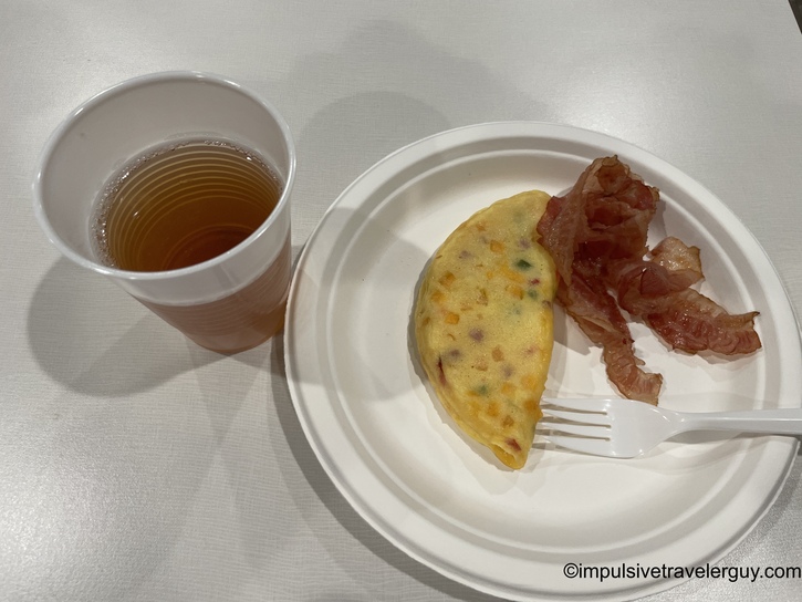 Hotel continental breakfast on white plates featuring vegetable omelet with three strips of bacon, plastic fork, and cup of coffee or tea on textured surface