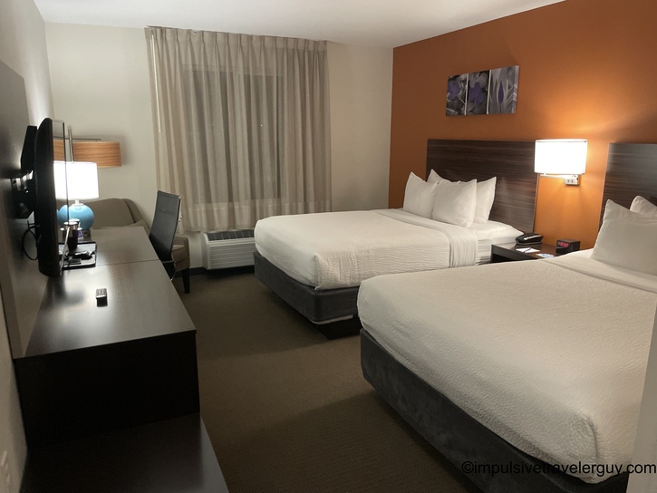 Clean hotel room with two queen beds, white linens, orange accent wall, desk area, flat-screen TV, and modern minimalist decor