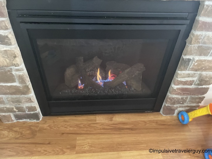 A gas fireplace with realistic ceramic logs and glowing flames, set into a brick surround with hardwood flooring visible at the base. A child&rsquo;s toy is partially visible in the corner.