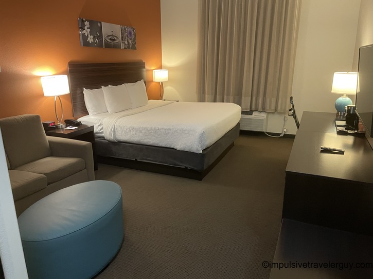 A hotel room interior with a king-size bed with white linens, orange accent wall, modern dark wood furniture, gray armchair with teal ottoman, and standard hotel amenities.