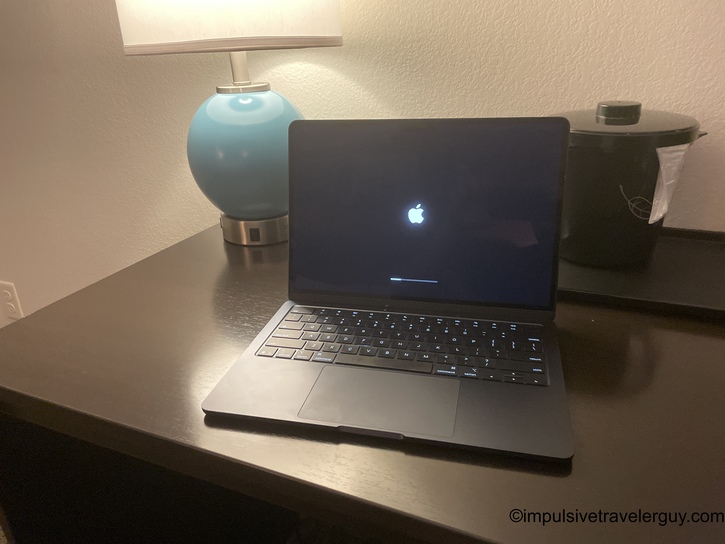 A MacBook laptop showing the Apple logo boot screen, sitting on a hotel room desk next to a teal spherical lamp and a small cooking pot.