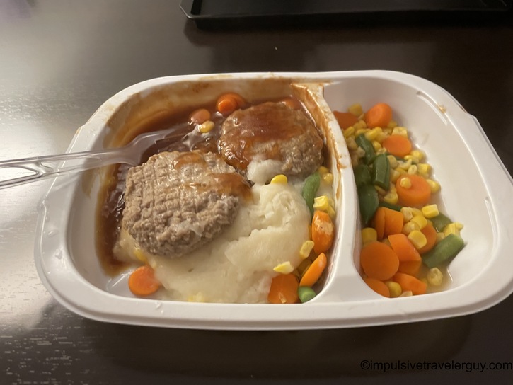 Cooked Hungry-Man salisbury steak frozen dinner in its white microwave tray, featuring two beef patties covered in brown gravy, mashed potatoes, and mixed vegetables including corn, carrots, and green beans. A plastic fork is visible on the side.
