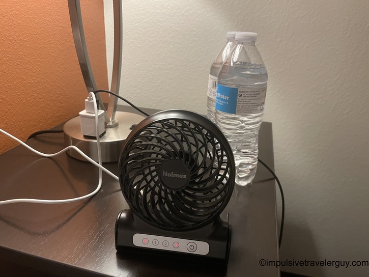 Hotel nightstand setup showing a small black Holmes brand portable desk fan, a water bottle, phone charger cables, and lamp base against an orange accent wall.
