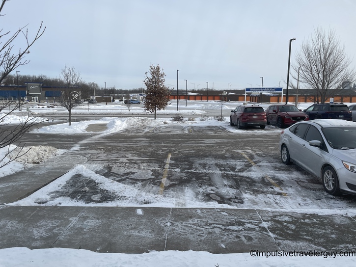 A winter parking lot scene with a Culver&rsquo;s restaurant visible in the background. The lot shows patches of ice and snow with slush, several parked vehicles, and overcast skies typical of Wisconsin winter weather.
