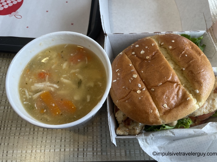 A Chick-fil-A meal consisting of a cup of chicken noodle soup with visible carrots and orzo pasta, alongside a grilled chicken sandwich on a multigrain bun with lettuce, served in the brand's signature packaging.