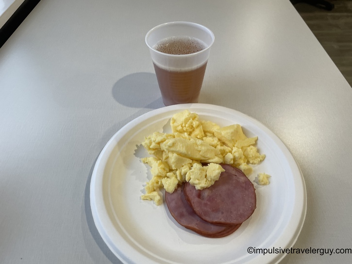 Hotel continental breakfast plate with scrambled eggs and two slices of Canadian bacon on a white styrofoam plate, accompanied by a plastic cup of apple juice on a white table.