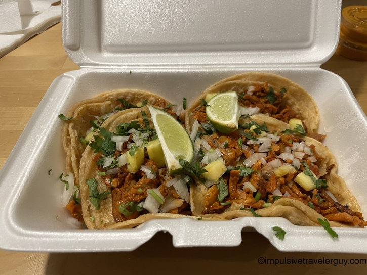 Three tacos al pastor served in a white styrofoam container, featuring seasoned pork topped with fresh cilantro, diced white onion, and pineapple chunks on corn tortillas, garnished with lime wedges.