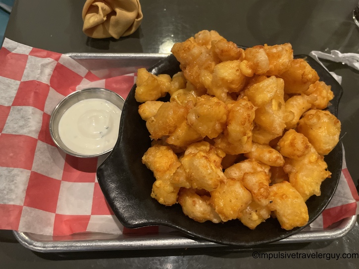 A generous portion of golden beer-battered Wisconsin cheese curds served in a black dish on red-and-white checkered paper, with a side of white ranch dipping sauce.