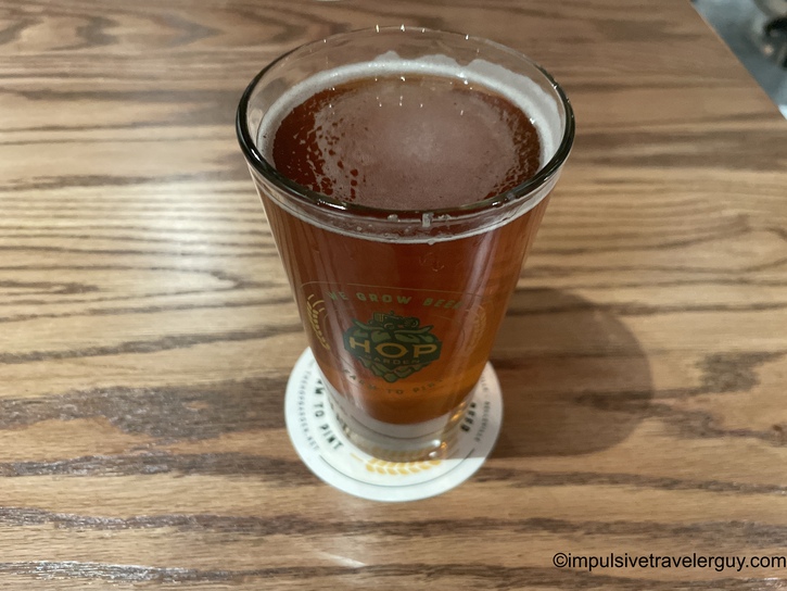 A pint glass of amber-colored craft beer with a foamy head, served in a glass featuring the Hop Garden logo, sitting on a wooden table.