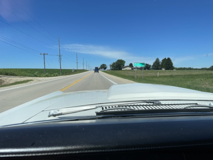 Behind the Cougar's wheel heading toward Dubuque, a green highway sign for Benton and Dubuque visible ahead, power lines running alongside flat farmland on both sides of a two-lane road.