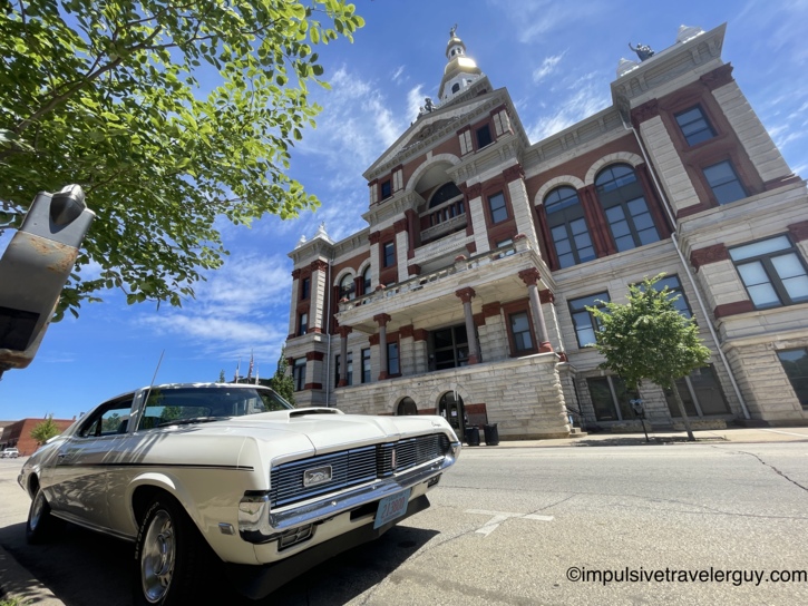 The white Cougar parked on the street in front of the Dubuque County Courthouse, a grand Romanesque Revival building in limestone and red brick with a gold-domed clock tower, arched windows, and ornamental stone carvings, shot from a low angle.